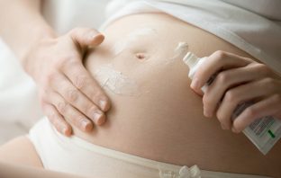 Close-up of torso of young pregnant model applying cream on her belly to prevent stretch marks. Future mom rubbing moisturizing cream on her tummy. Pregnancy skincare concept